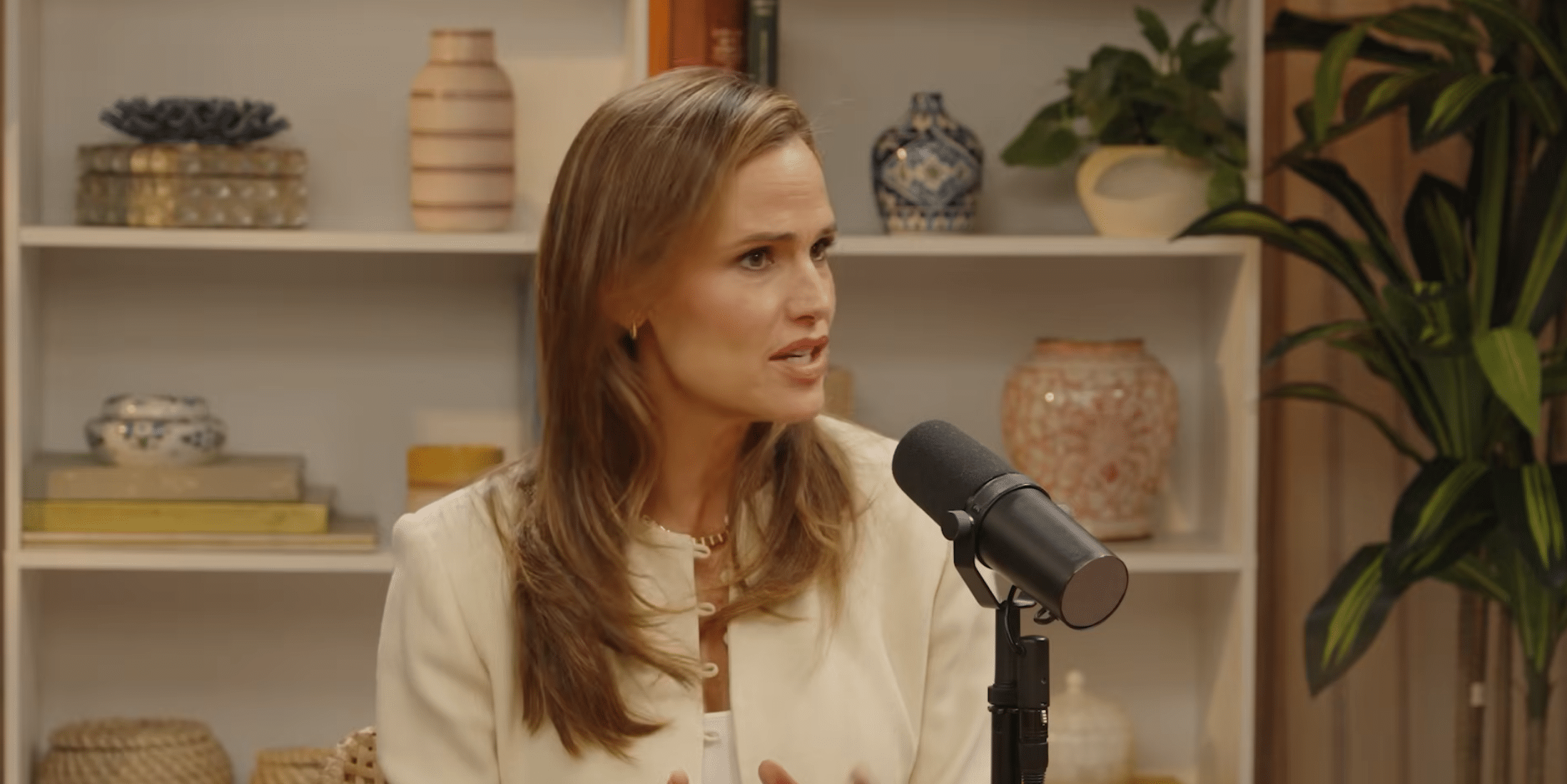 A woman with long light brown hair sits speaking into a microphone, clearly engaged in video production. She wears a white blazer and a gold necklace. Behind her are shelves with potted plants, books, and decorative vases.